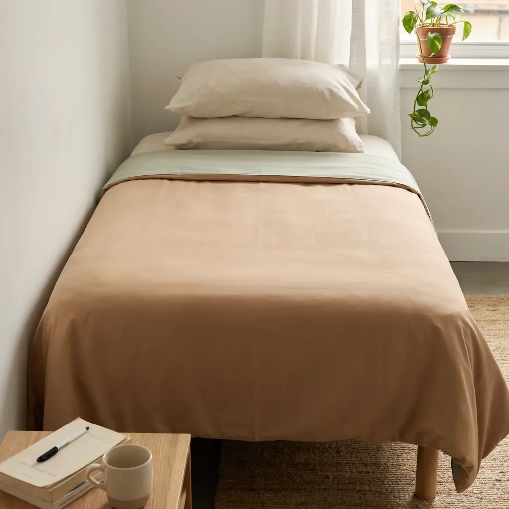 Reversible twin XL dorm comforter showing warm sand folded back to reveal muted sage green reverse with cream pillows
